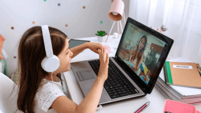 A young girl wearing headphones sits at a desk and waves at a woman on a video call displayed on her laptop.