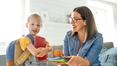 A woman hands a young girl notebooks and an apple. The girl has a backpack and holds a red book while smiling. They are indoors in a brightly lit room.