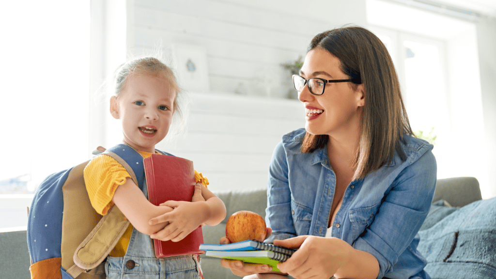A woman hands a young girl notebooks and an apple. The girl has a backpack and holds a red book while smiling. They are indoors in a brightly lit room.