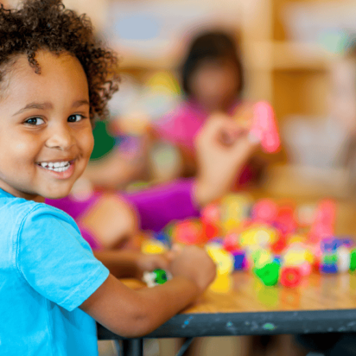 Smiling child in a blue shirt sits at a table with colorful building blocks, with other children playing in the blurred background.
