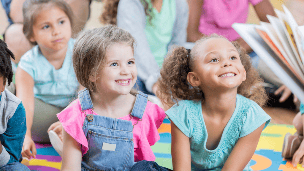 Several young children sit on a colorful mat, smiling and looking at an unseen person holding a book, suggesting they are engaged in a group storytime or classroom activity.