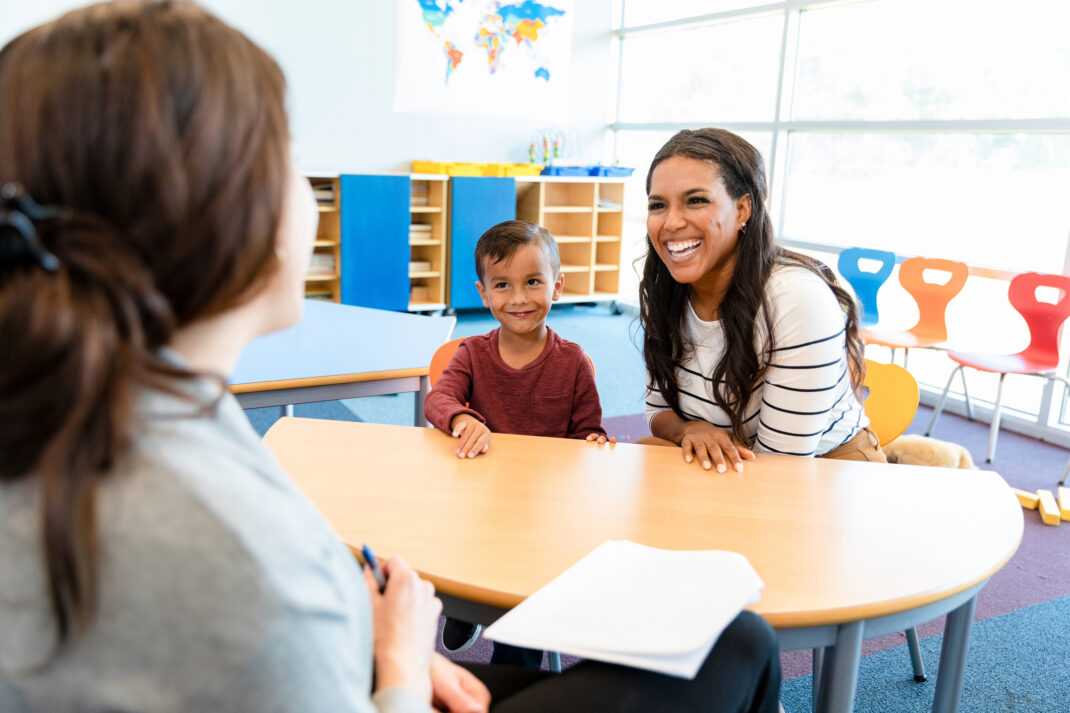 A woman and a young boy sit at a table smiling at another woman, possibly a teacher, in a brightly lit classroom setting.