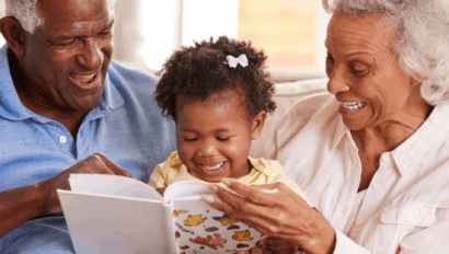 An elderly man and woman sit on a couch with a young girl, smiling as they read a picture book together.