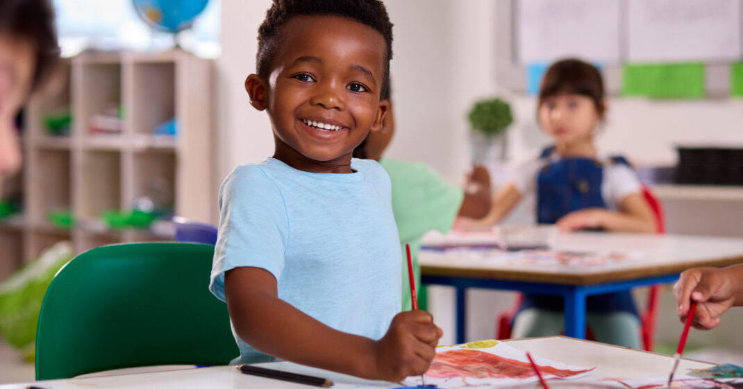 Smiling young boy in a light blue shirt sits at a classroom table, holding a paintbrush and working on an art project. Other children are visible in the background.